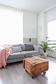 Pink blanket and various scatter cushions on pale grey couch behind vintage wooden crate used as coffee table