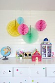 Colourful paper pompoms hung on white wood cladding above toys on chest of drawers in girl's bedroom