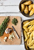 Ingredients for rosemary potatoes on a wooden chopping board and raw potato wedges in a roasting dish
