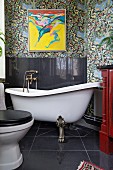 Vintage Badezimmer mit freistehender Badewanne auf Löwenfüssen, schwarz gefliester Spritzschutz an Wand, oberhalb modernes Bild auf floraler Tapete