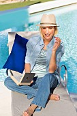 Blonde Frau in Jeans, Bluse, Ringelshirt und Hut sitzt an Pool und liest