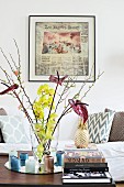 Glasvase mit Blüten, Kerzen und Bücherstapel vor gerahmtem Zeitungsausschnitt und Couch