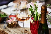 A glass of rosé champagne, a bunch of spring flowers and a bottle of champagne on a wooden table