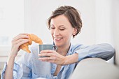 Frau sitzt entspannt mit Croissant und Kaffetasse auf Couch