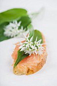 A slice of baguette topped with smoked salmon, a wild garlic leaf and edible wild garlic flowers