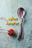 The words 'Guten Appetit' spelt with alphabet pasta next to cutlery and a tomato