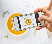 A woman using a smartphone to take a picture of carrot and pumpkin soup