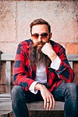 A man with a beard, sunglasses and a lumberjack shirt sitting on a bench
