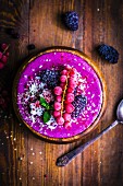 A smoothie bowl with frozen redcurrants, blackberries, dragon fruit flowers and coconut on a wooden surface