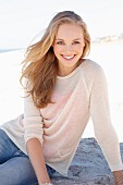 Dunkelblonde junge Frau in hellem Strickpulli und Jeans am Strand