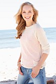 A young dark blonde woman on a beach wearing a light knitted jumper and jeans