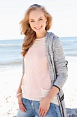 A young dark blonde woman on a beach wearing a light knitted jumper, a cardigan and jeans