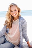 A young dark blonde woman on a beach wearing a light knitted jumper, a cardigan and jeans