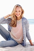 Dunkelblonde junge Frau in hellem Strickpulli, Strickjacke und Jeans am Strand