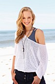 A young blonde woman on a beach wearing a tank top and a net jumper