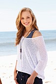 A young blonde woman on a beach wearing a tank top and a net jumper