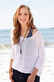 A young blonde woman on a beach wearing a tank top and a net jumper