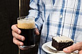 A man holding a slice of chocolate cake and a glass of dark beer