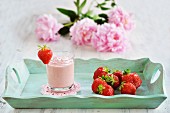 A glass of strawberry yoghurt and fresh strawberries on a wooden tray with a bunch of fresh summer flowers in the background