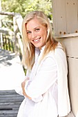 A young blonde woman leaning on a wooden facade wearing a white blouse