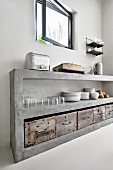 Beton-Küchenregal mit Geschirrstapeln, Gläsern und rustikalen Holzkisten unter geöffnetem Fenster
