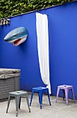 Colourful metal stools in front of shark's head and curtain on blue garden wall