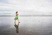 Frau in grünem Sommerkleid geht mit Handy in der Hand am Strand entlang