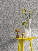 Gelber Metalltisch mit Vasen vor Vliestapete mit floralem Muster in Grautönen