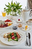 A breakfast table with prosciutto, mixed leaves, balsamic vinegar, bread and butter, cucumber, radishes, a soft boiled egg and a cheese plate