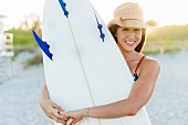 A young American woman with a cowboy look holding a surfboard on the beach
