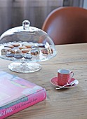 Cake stand under glass cover, cup and book on wooden table