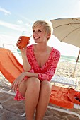A blonde woman wearing a patterned tunic dress sitting on a sunlounger and drinking a smoothie