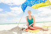 Blonde Frau in türkisfarbenem Top sitzt auf Badetuch am Strand