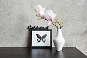 Magnolienblüte in weisser Vase vor Bild mit Schmetterlingsmotiv an Betonwand