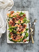 Prosciutto, melon, figs and soft cheese on a white serving board over grunge background