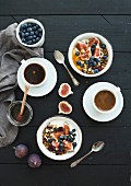 Bowls of oat granola with yogurt, fresh blueberries and figs, cups of coffee and honey