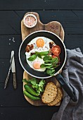 Pan of fried eggs, bacon, cherry tomatoes and fresh herbs with bread on wooden board over dark wooden background