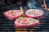 Fresh steaks seasoned with pepper and rosemary on a grill rack with fire