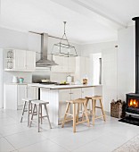 Black log-burning stove and rustic wooden bar stools at counter in white, open-plan kitchen