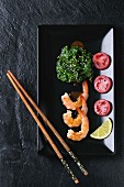 Spinach with sesame seeds, pan-fried prawns, lemon and tomato on a black serving dish with chopsticks