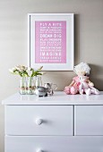 Picture with saying above the chest of drawers with flowers and dolls