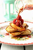 Maple syrup being poured over pancakes with raspberries