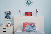 Kinderzimmer mit weißem Bett und Nachtschränkchen vor hellblauer Wand dazu rote Farbakzente durch Kissen, Lampen und Deko