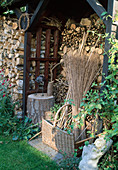 Gartenhäuschen mit Brennholz-Stapel, Frauentorso, Metallvogel auf Hackstock, Korb mit Grasbündeln und Bambusstangen