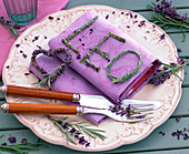Lavandula (Lavendel) zu Herz und Buchstaben 'Leo' gebunden auf lila Serviette