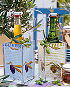 Flaschen mit Olivenöl in Tüten mit Oliven-Motiven, Zweige und Früchte von Olea