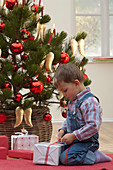 Kleiner Junge mit Geschenk sitzt vor Weihnachtsbaum