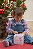 Kleiner Junge mit Geschenk sitzt vor Weihnachtsbaum