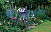 COVERED SEAT PAINTED Mauve SURROUNDED by Metal URNS PAINTED Silver with AGAVES, Silver Metal OBELISKS, ALLIUM CHRISTOPHII, HEMEROCALLIS, CARDOON AND ERIGERON. THE NICHOLS Garden