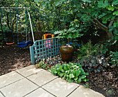 CHILDRENS Garden: SWINGS with BARK BENEATH, Blue TRELLIS Screen, Water Feature with FATSIA JAPONICA BEHIND. Designer: Sarah LAYTON